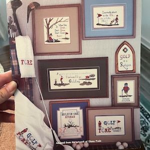 May include: A book cover titled "GOLF-ISMS" featuring framed cross-stitch artworks with golf-themed sayings and imagery. Includes a golf towel, golf clubs, and golf balls. The text includes "FORE" and "GOLF is a better word!"