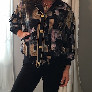 Puede incluir: Una chaqueta bomber de satén negro con ribetes dorados y un estampado abstracto colorido. La chaqueta tiene cierre de botones y cuello.