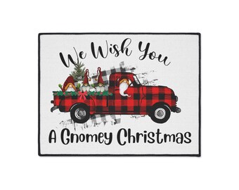 Può includere: Un zerbino natalizio rettangolare con bordo nero. Il design presenta un camion a scacchi rossi e neri che trasporta gnomi e un albero di Natale. Il testo recita "We Wish You A Gnomey Christmas."