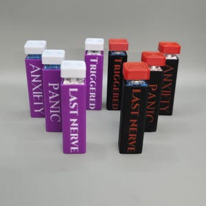 May include: An assortment of rectangular, labeled containers. Some are purple with white text reading "ANXIETY", "PANIC", and "LAST NERVE". Others are black with red text reading "TRIGGERED" and "LAST NERVE". All have colored caps.