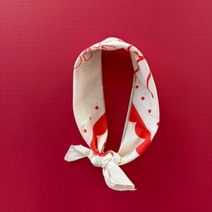 Puede incluir: Bandana color crema con un patr&oacute;n rojo de puntos y formas curvas. La bandana est&aacute; anudada en la parte inferior y descansa sobre un fondo rojo liso. La tela parece ligera.