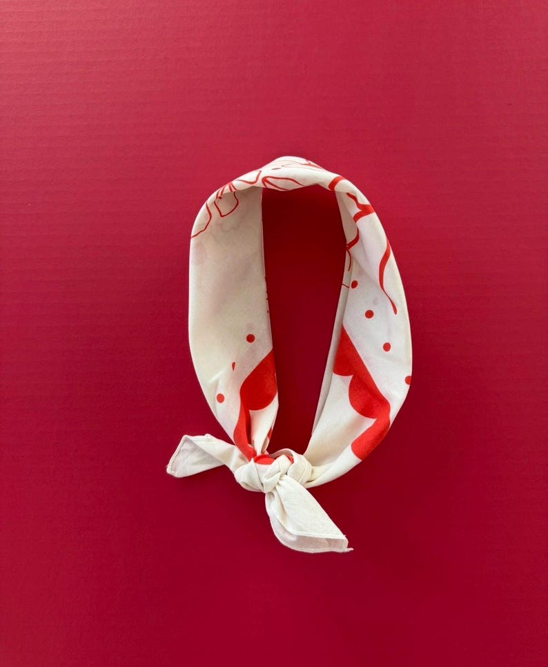 Puede incluir: Bandana color crema con un patr&oacute;n rojo de puntos y formas curvas. La bandana est&aacute; anudada en la parte inferior y descansa sobre un fondo rojo liso. La tela parece ligera.