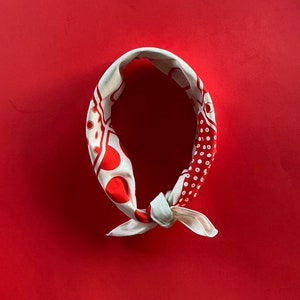 May include: A cream-colored bandana with a red polka dot and geometric pattern. The bandana is tied in a knot and lies on a red background. The design features large and small red circles and other shapes.