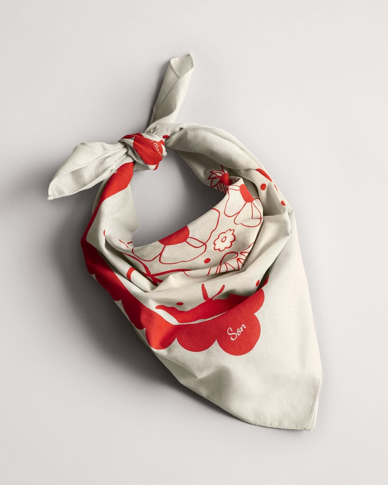 Puede incluir: Un pa&ntilde;uelo de color crema con un estampado floral rojo y la palabra "Son" impresa. El pa&ntilde;uelo est&aacute; anudado en la parte superior, mostrando el dise&ntilde;o. La tela est&aacute; doblada, creando una forma triangular.