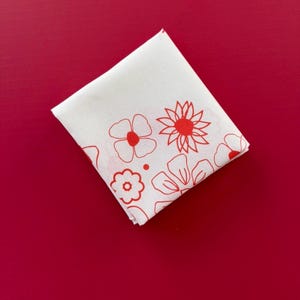 Puede incluir: Pa&ntilde;uelo cuadrado blanco con un dise&ntilde;o floral rojo. Las flores est&aacute;n delineadas en rojo y tienen centros rojos. El pa&ntilde;uelo est&aacute; doblado y colocado sobre un fondo rojo liso.