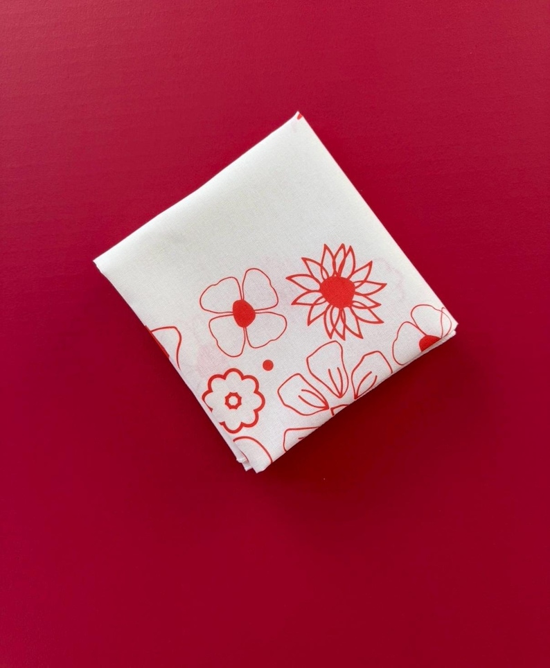 Puede incluir: Pa&ntilde;uelo cuadrado blanco con un dise&ntilde;o floral rojo. Las flores est&aacute;n delineadas en rojo y tienen centros rojos. El pa&ntilde;uelo est&aacute; doblado y colocado sobre un fondo rojo liso.