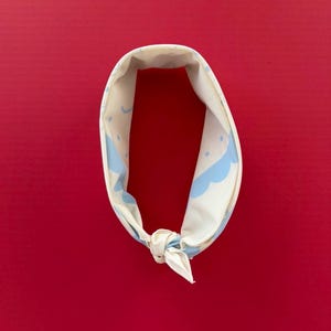 Puede incluir: Bandana color crema con un patr&oacute;n repetido de nubes y puntos azul claro. La bandana est&aacute; atada con un nudo en la parte inferior y se muestra sobre un fondo rojo.