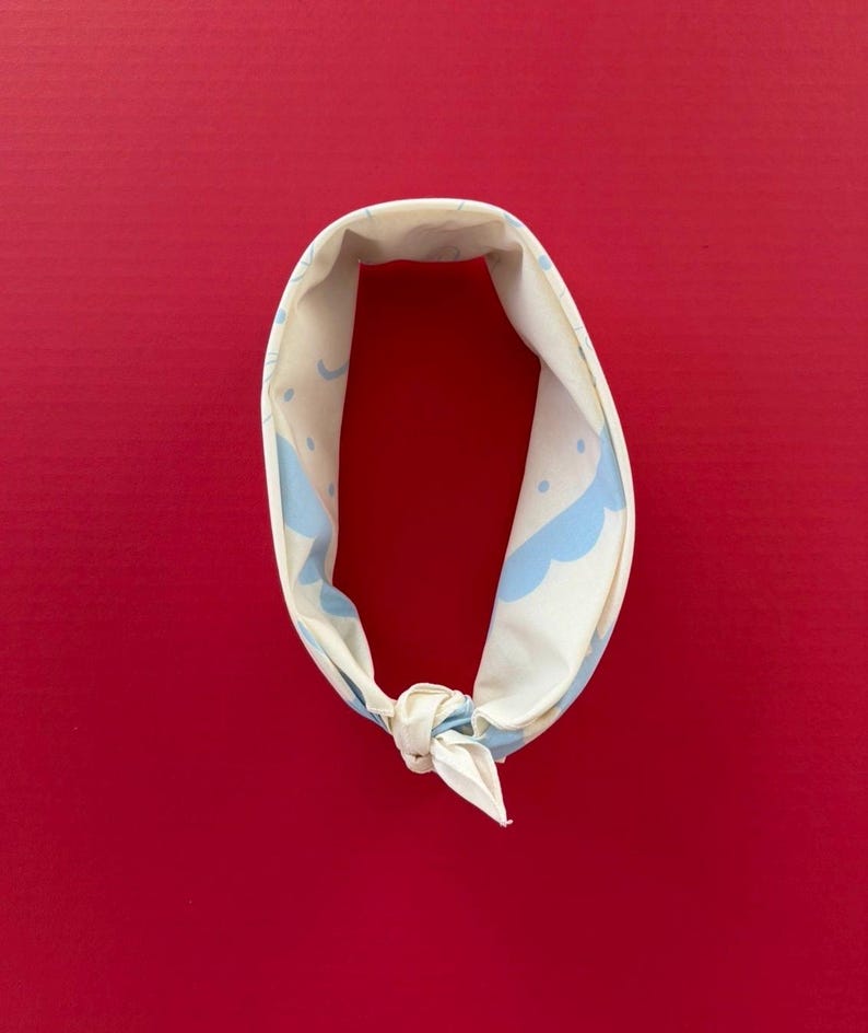 Puede incluir: Bandana color crema con un patr&oacute;n repetido de nubes y puntos azul claro. La bandana est&aacute; atada con un nudo en la parte inferior y se muestra sobre un fondo rojo.