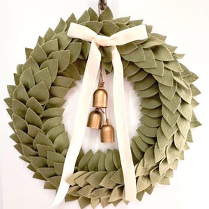 May include: A circular wreath constructed from layered olive green felt leaves. A cream-colored velvet ribbon is tied at the top, with two long tails hanging down. Three small, metallic gold-toned bells are suspended from the center of the ribbon.