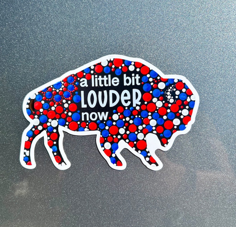 May include: A black and white buffalo silhouette with red, white, and blue polka dots. The buffalo has the text "a little bit LOUDER now" written on it.