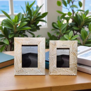 May include: Two decorative photo frames with a cream and gold swirling pattern. The frames have a rectangular shape with a central opening for a photo. The frames are displayed on a wooden surface with greenery in the background.