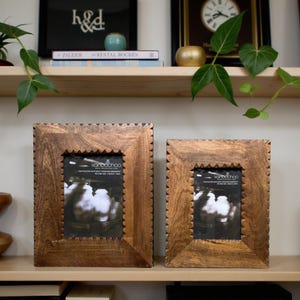 May include: Two square wooden photo frames with a dark brown finish and a decorative edge. The frames are displayed on a wooden shelf. The frames have a natural wood grain and a decorative edge.