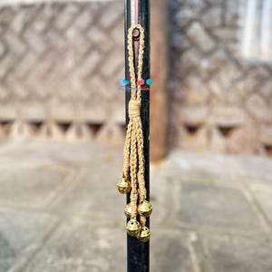 May include: A jute tassel with six small, gold-coloured bells. The tassel is attached to a loop and hangs from a black metal pole. The background is a blurred outdoor scene with stone steps.