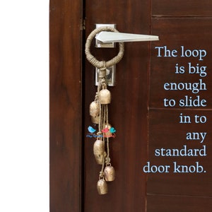 May include: A brown rope door knocker with a loop large enough to fit over a standard door knob. The knocker has a string of small, gold-coloured bells hanging from it. The text "The loop is big enough to slide in to any standard door knob." is written in blue text on the right side of the image.