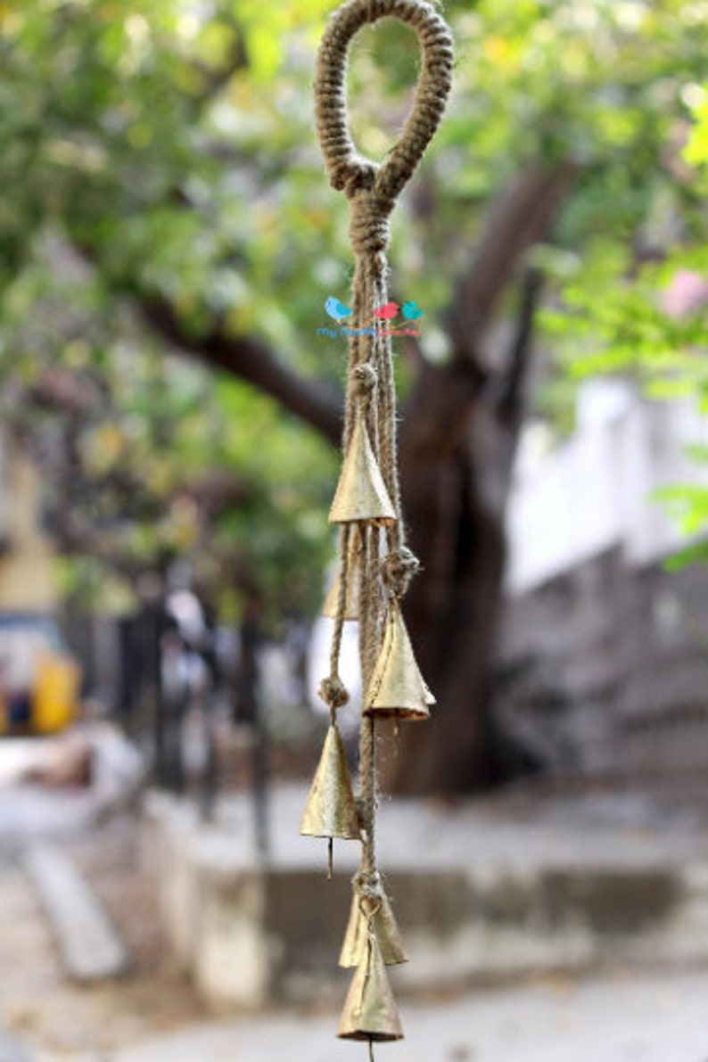 May include: A gold wind chime with a jute rope and multiple bells hanging from it. The chime is hanging from a loop of jute rope.