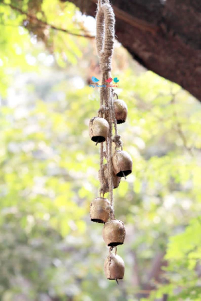 May include: A string of small, gold-coloured bells hanging from a thick, natural rope. The bells are arranged in a cascading pattern, with the largest bell at the bottom. The rope is tied in a loop at the top, suggesting it can be hung from a ceiling or branch.