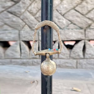 Op de afbeelding: Een messing bel met een touwhandvat bevestigd aan een zwarte paal. De bel heeft een rond gezicht met een inscriptie in een vreemde taal.