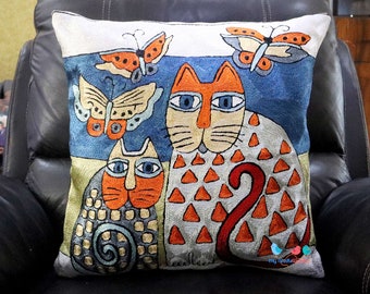 Peut inclure: Coussin carré brodé avec deux chats stylisés et trois papillons sur un fond bleu et jaune. Les chats ont des détails orange et gris, le plus grand chat ayant des motifs triangulaires orange. Les papillons sont orange et crème.