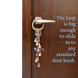 May include: A brown wooden door with a silver door knob. A jute rope door knocker with a loop and a string of small gold bells hangs from the door knob. The text "The loop is big enough to slide in to any standard door knob." is written in blue text on the right side of the image.