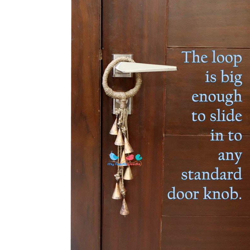 May include: A brown wooden door with a silver door knob. A jute rope door knocker with a loop and a string of small gold bells hangs from the door knob. The text "The loop is big enough to slide in to any standard door knob." is written in blue text on the right side of the image.
