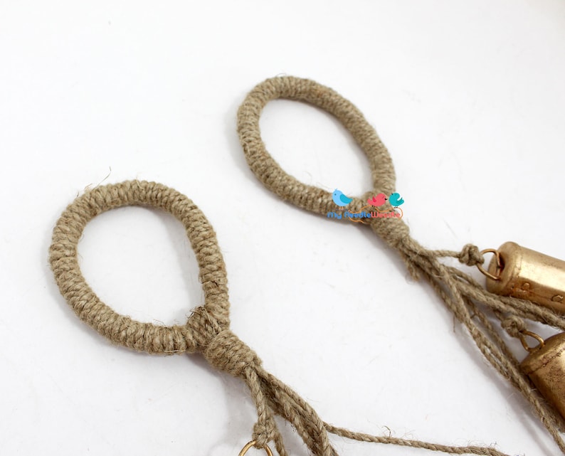 May include: Two brown jute rope loops with gold bells hanging from the bottom. The loops are tied together with a knot.