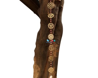 Op de afbeelding: Een gouden metalen chakra-wandhanger met zeven verschillende chakra-symbolen. De symbolen zijn verbonden door een rode kralenketting. De hanger is bevestigd aan een boomstam.