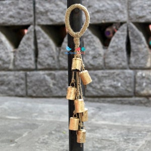 May include: A decorative hanging with a loop of natural twine and a series of small gold bells attached to it. The bells are hanging from the twine and are arranged in a cascading pattern.
