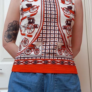 May include: A sleeveless cream-coloured top featuring a repeating print of red and black horse-drawn carriages. The centre panel displays a dark brown grid pattern with small orange dots, finished with a solid orange hem and a decorative border.