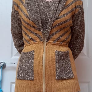 May include: Mustard yellow knitted cardigan with a zip-up front, hood, and two front pockets. The cardigan features brown striped accents on the sleeves and hood. The waist is cinched with a knitted band. The model is wearing blue jeans.