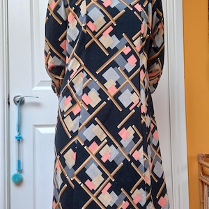 May include: A knee-length dress with long sleeves, featuring a geometric pattern. The dress has a black base with a repeating design of squares and lines in grey, peach, and tan. It has a round neckline and a simple, classic cut.