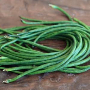 May include: A pile of fresh, vibrant green long beans, also known as yardlong beans, on a dark brown wooden surface. The beans are long, slender, and slightly curved, with a smooth texture. The image highlights the natural color and shape of the vegetables.