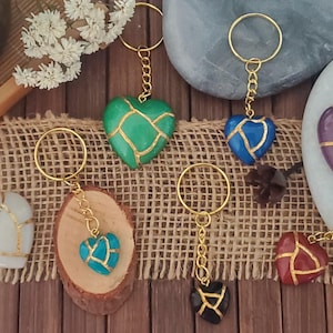 Può includere: Otto portachiavi a forma di cuore dorati con diversi colori di resina e dettagli dorati. I cuori sono crepati e hanno linee dorate che li attraversano. I portachiavi sono su una superficie di legno marrone.