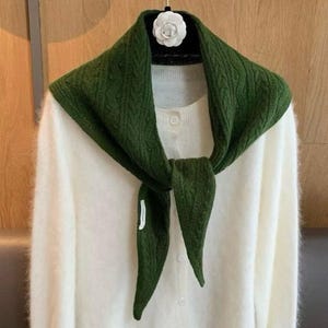 May include: A white long-sleeved fuzzy cardigan with a button-down front, paired with a dark green cable-knit triangular scarf draped around the neck. The scarf features a small white rectangular tag on one end and a white flower-shaped pin.