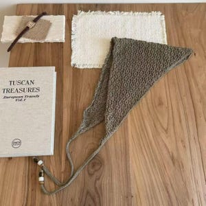 May include: A still-life arrangement featuring a crochet scarf, a book titled "Tuscan Treasures," a watch, and a small woven mat. The scarf is olive green, and the book has the text "European Travels Vol. 1." The watch has a brown leather strap.