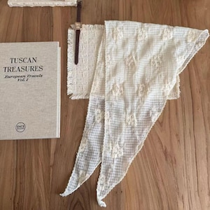 Puede incluir: Una bufanda color crema con bordados florales, un libro titulado "Tuscan Treasures European Travels Vol. I" y un reloj de cuero marrón con esfera rectangular se exhiben sobre una superficie de madera.