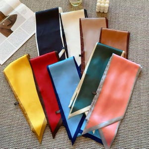 May include: A collection of long, rectangular scarves in various solid colors including yellow, red, light blue, dark green, peach, brown, beige, white, and black. Each scarf features a smooth, satin-like finish and a pointed, angled end.
