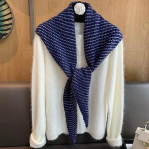 Pode incluir: Um suéter branco de manga comprida com textura felpuda, apresentando uma frente abotoada e um enfeite de flor branca na gola. Um lenço triangular com padrão azul-escuro e branco está amarrado ao redor do pescoço sobre o suéter.