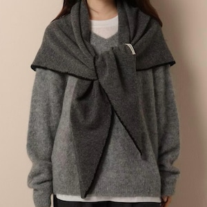 May include: A dark charcoal grey knit scarf draped over a light grey long-sleeve sweater. The scarf features a small white rectangular fabric tag on the side. The sweater has a V-neckline and is layered over a plain white undershirt.