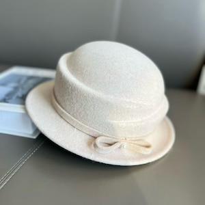 May include: A cream-coloured cloche hat with a rounded crown and a small brim. The hat features a decorative ribbon with a bow and a small pearl accent. The hat is made of a soft, felt-like material.