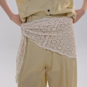 May include: Cream-colored, crocheted belt or scarf draped over light beige pants. The belt features a floral pattern and is tied at the waist. The person is wearing a light yellow button-down shirt.