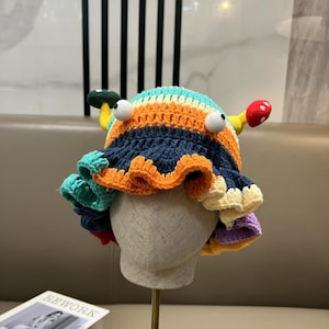 Puede incluir: Un sombrero de ganchillo hecho a mano con diseño de cangrejo. El sombrero presenta una corona a rayas en tonos azules, naranjas y amarillos, con ojos grandes y ondulados y bordes coloridos con volantes. El sombrero tiene una estética lúdica y única.