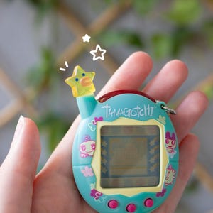 Op de afbeelding: Een talingkleurig Tamagotchi digitaal huisdierapparaat met een gele stervormige antenne, een scherm met een pixelweergave en drie roze ronde knoppen. De behuizing is versierd met kleurrijke karakterafbeeldingen en het woord Tamagotchi in het wit.