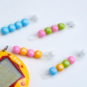Op de afbeelding: Een geel draagbaar elektronisch spel met een scherm, naast verschillende kleurrijke kralenaccessoires. De kralen zijn blauw, groen, roze en oranje, elk met de letters "TMGC" erop gedrukt. De accessoires hebben witte lussen.