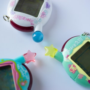 Peut inclure: Trois appareils de compagnie numérique Tamagotchi avec des coques en plastique ovales blanches, bleu sarcelle et bleu clair. Chaque appareil possède un petit écran, une antenne saillante avec un embout en forme d'étoile ou de sphère et le logo Tamagotchi.