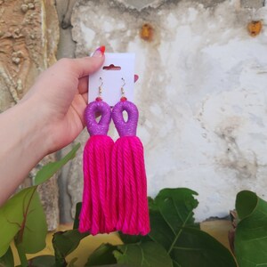Peut inclure: Une paire de boucles d'oreilles en pompon rose avec des accents violets et argent&eacute;s. Les boucles d'oreilles sont sur une carte blanche.