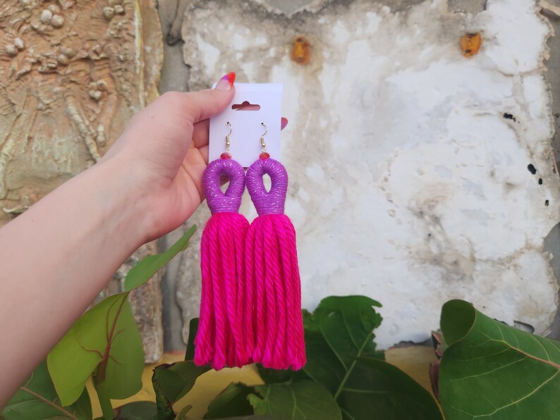 Peut inclure: Une paire de boucles d'oreilles en pompon rose avec des accents violets et argent&eacute;s. Les boucles d'oreilles sont sur une carte blanche.