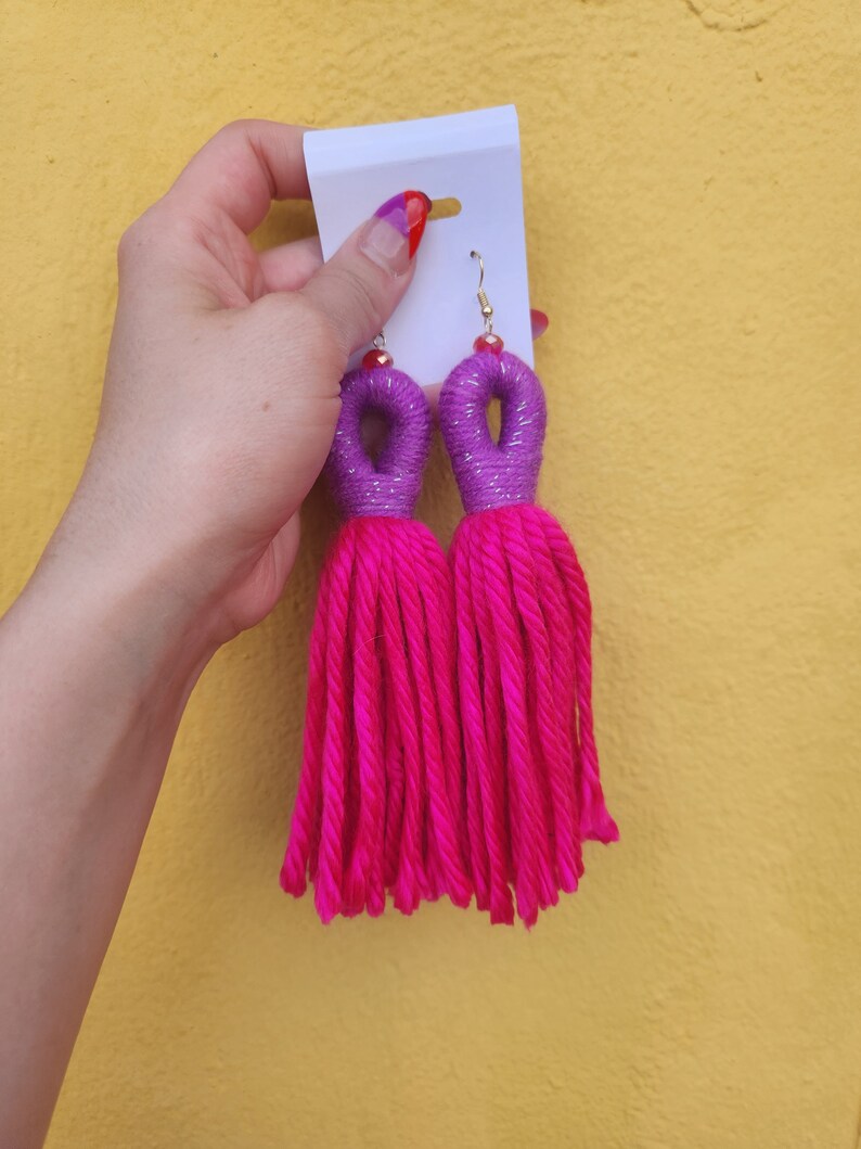 Peut inclure: Une paire de boucles d'oreilles en pompon rose avec des accents violets et argent&eacute;s. Les boucles d'oreilles sont faites &agrave; la main et pr&eacute;sentent un design unique.