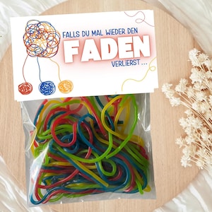 Könnte beinhalten: Eine weiße Karte mit dem Text "Falls du mal wieder den Faden verlierst..." und einer bunten Illustration von verwickeltem Garn. Die Karte ist an einem durchsichtigen Plastikbeutel befestigt, der mit bunten Lakritzschnüren gefüllt ist.