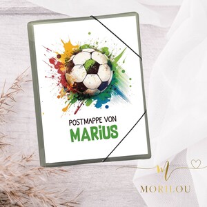 Könnte beinhalten: Ein grauer Ordner mit weißem Hintergrund und einer bunten Fußballgrafik. Der Text "Postmappe von Marius" ist auf dem Ordner gedruckt.