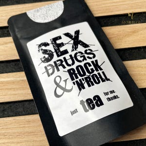 Könnte beinhalten: Schwarzer Teebeutel mit weißem Etikett. Das Etikett zeigt den durchgestrichenen Text "SEX DRUGS & ROCK 'N' ROLL" mit "tea" und "just for me, thanks." darunter. Der Teebeutel hat eine abgerundete Oberseite.